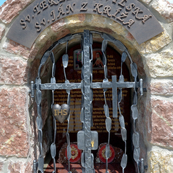 Schmiedeeisernes Denkmal der Heiligen mit Attributen auf dem Gitter. Herz mit der Inschrift IHS und Kreuz